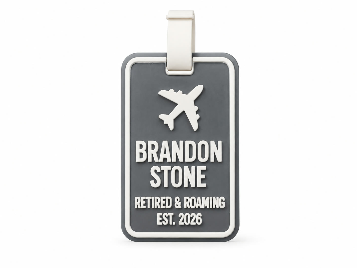 A studio render on white of a dark grey 3D printed luggage tag with a raised white border, a raised white aeroplane and the raised white lettering BRANDON STONE, RETIRED & ROAMING, EST. 2026, with a white flexible strap looped through the slot at the top.