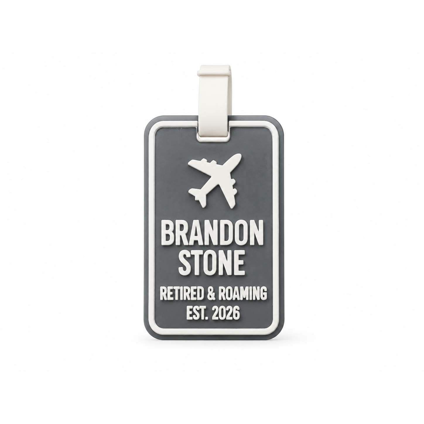 A dark grey 3D printed luggage tag with a raised white border, a raised white aeroplane, and raised white lettering reading BRANDON STONE, RETIRED & ROAMING, EST. 2026, with a white flexible strap looped through the top.