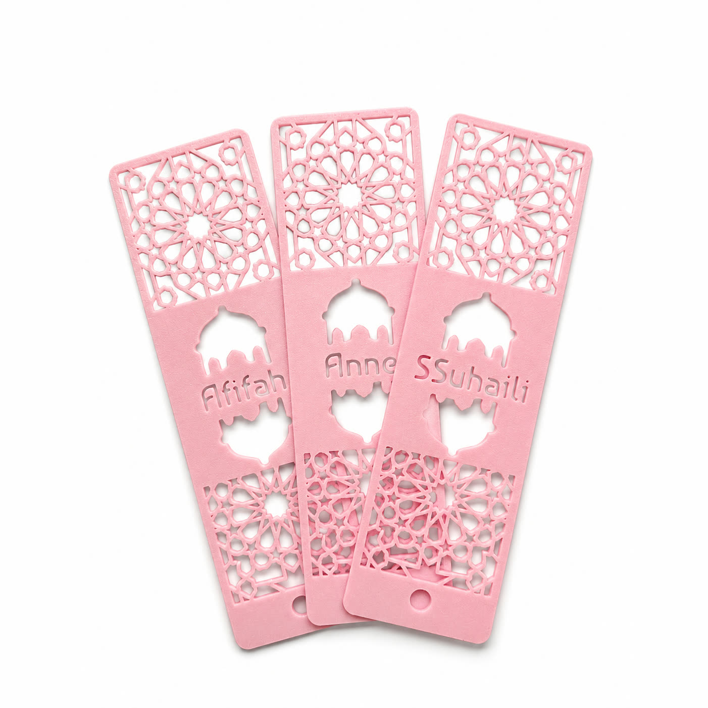 Three pink 3D printed bookmarks fanned out, each with a name set inside a cut-out mosque arch, with geometric lattice panels above and below.